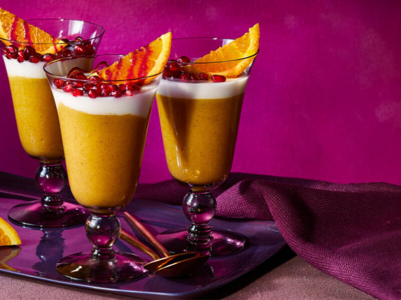 Jamaican Orange Cornmeal Pudding with Coconut Fluff in parfait glasses served with a wedge of orange and pomegranate seeds