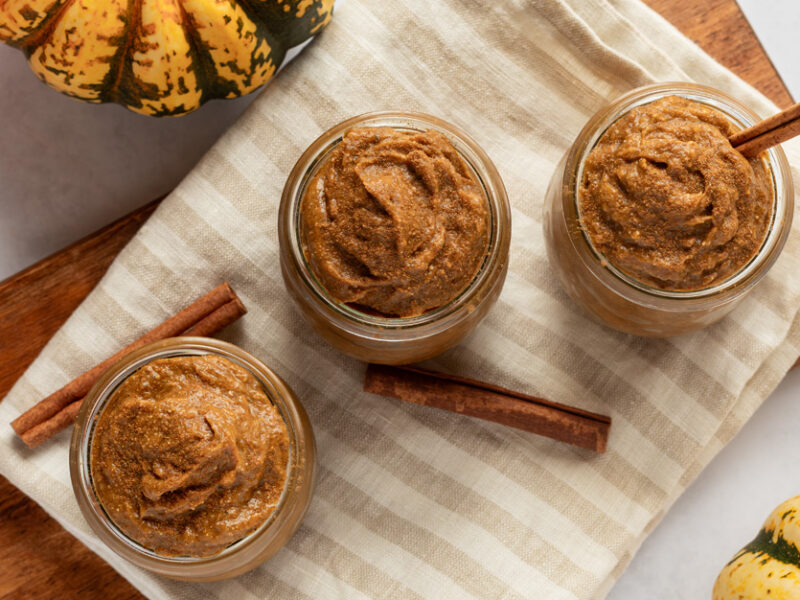 A top-down shot of three jars of almost-instant pumpkin pudding, with two sticks of cinnamon and some pumpkins lying beside the glasses, an easy vegan dessert recipe by Chef AJ