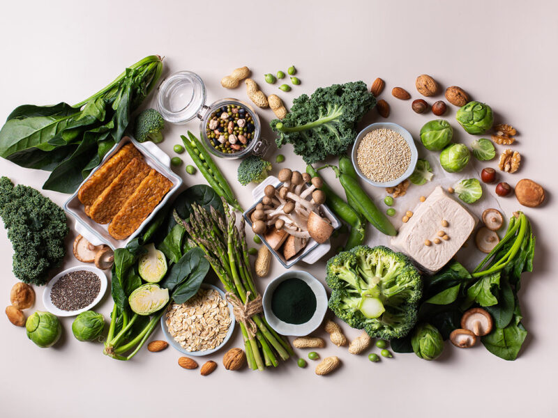 a wide array of protein-rich whole plant foods shown arranged on a white surface, including tofu, broccoli, tempeh, nuts, and seeds