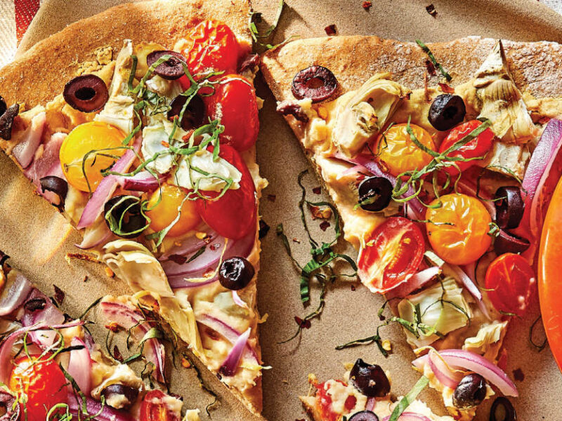 Mediterranean Artichoke Pizza on a light-weight wooden platter next to a small red bowl of lemons, with a red and white striped dish towel behind