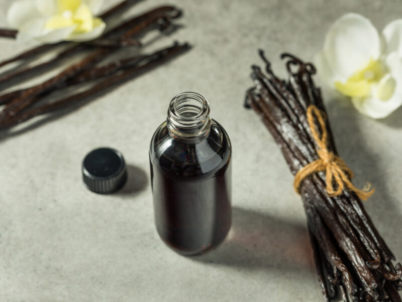 A bottle of vanilla extract beside whole vanilla bean pods and a flower