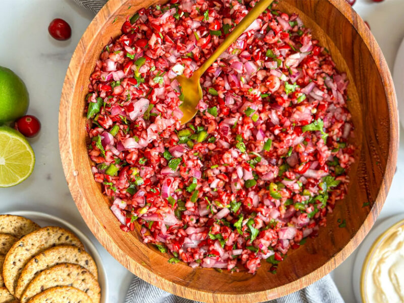 Jackie Akerberg’s Cranberry Salsa in a wooden serving bowl with a copper spoon, beside limes and crackers and vegan ricotta