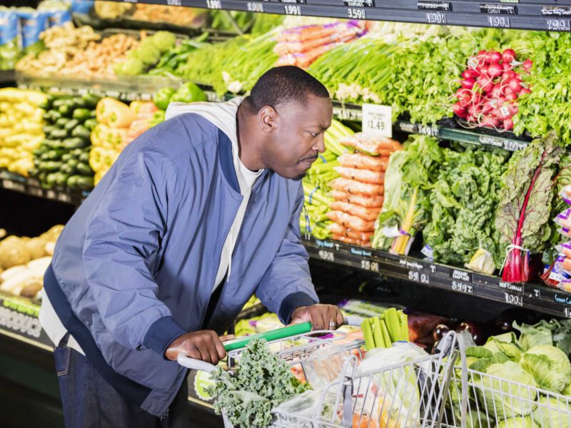 A Black man in his 40s shops the produce section of a grocery store, with a cart full of vegetables