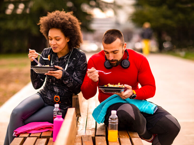 Two vegan athletes, a woman and man who appear to be in their early 30s, sit side by side on benches eating a healthy lunch