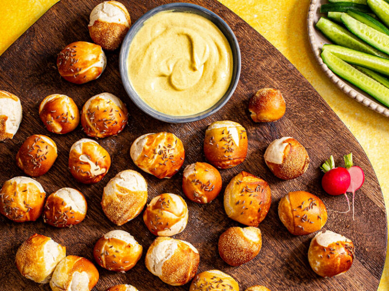 Whole Grain Pretzel Bites on a platter with mustard next to plates of sliced cucumber, radish, and pickles
