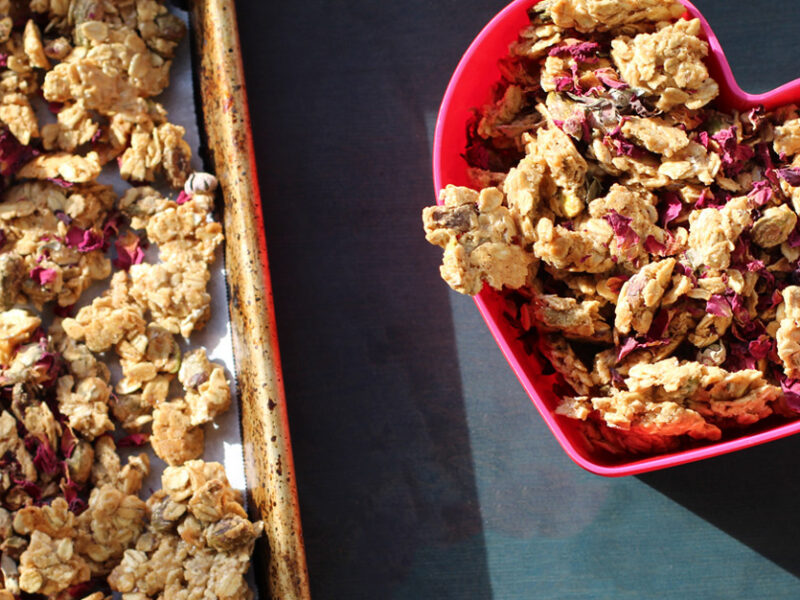 A ceramic baking dish filled with Rose Tahini Granola sits on a dark grey counter, next to a pink heart-shaped bowl of the granola