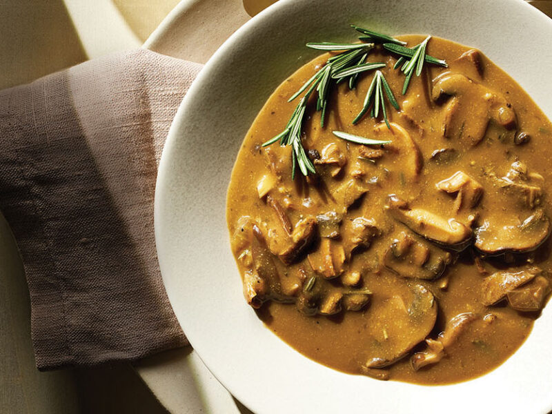A creamy-looking bowl of Wild Mushroom Soup with a sprig of fresh rosemary on top. A brown cloth napkin sits under the white bowl.
