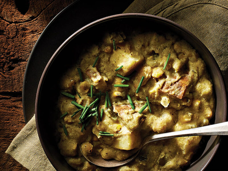 A dark grey ceramic bowl of Dairy-Free Potato Corn Chowder in a cozy, dimly lit setting