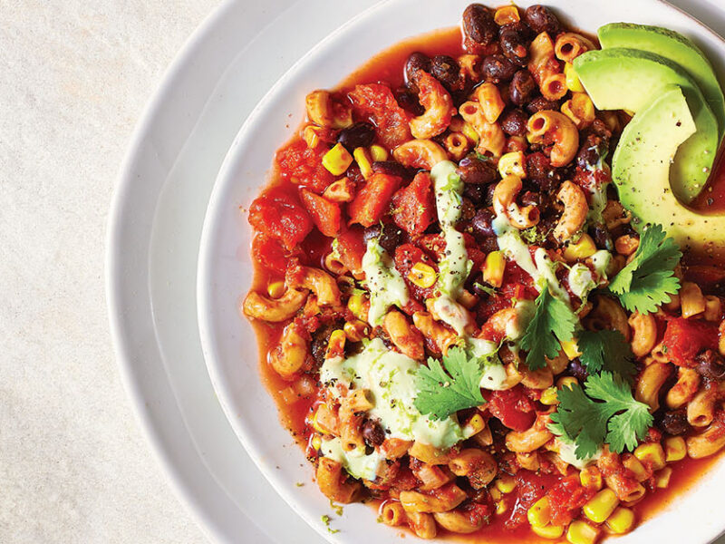 A white, wide-rimmed bowl of Zesty Macaroni with Black Beans, Corn, and Avocado Cream, nested on a white plate on a white table cloth