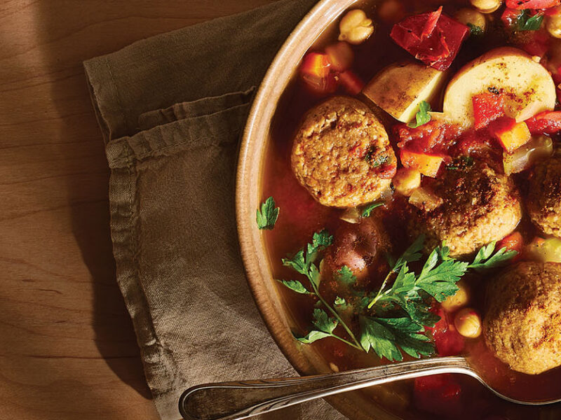 A rustic brown serving bowl of Vegan Kefta Tagine (Moroccan Meatballs) on a brown cloth napkin, with a spoon in the bowl