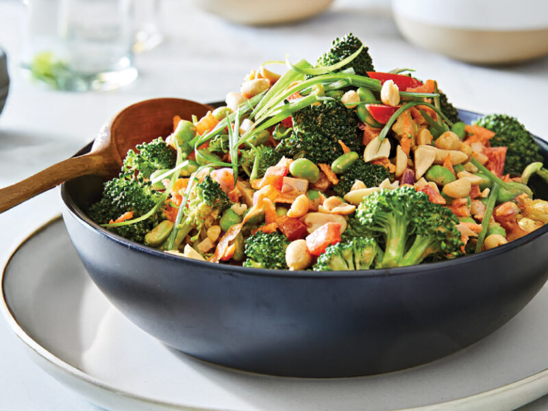 Broccoli Crunch Salad in a black bowl against a white background, with a wooden spoon in the bowl