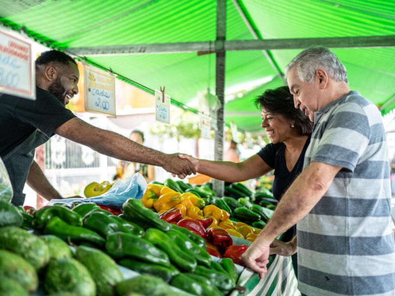An older couple shops at a farmers market, looking at bell peppers, and the woman shakes the vendor's hand as they smile