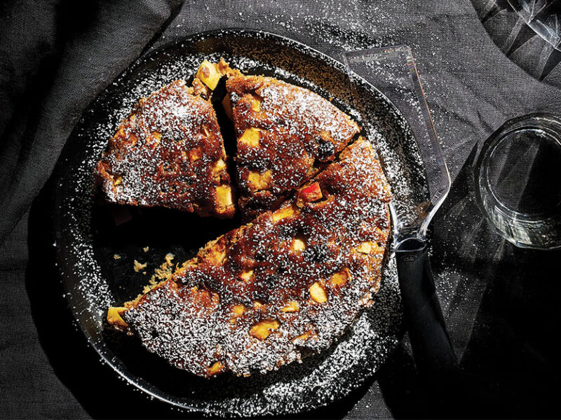 Top-down view of Air-Fryer Apple Fritter Cake dusted with powdered sugar, on a grey tablecloth next to a class of water