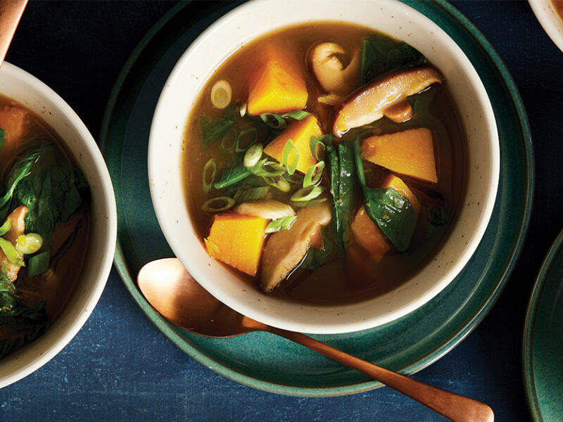 Four bowls of Thai Lemongrass Soup with Mushrooms, Squash, and Greens, on a dark blue tablecloth, with a spoon in each bowl