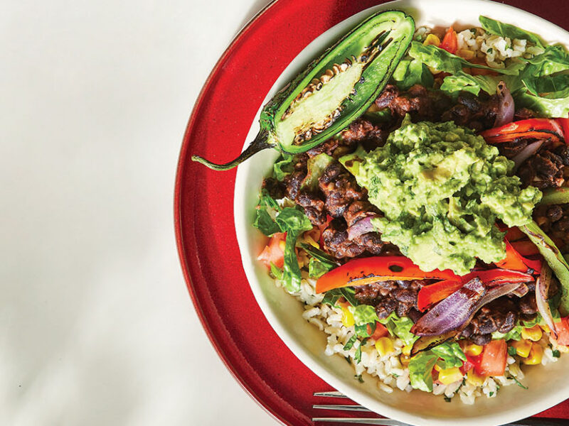Veggie Burrito Bowl topped with guacamole, cooked red onion and red bell pepper, in a white bowl nested on a red plate