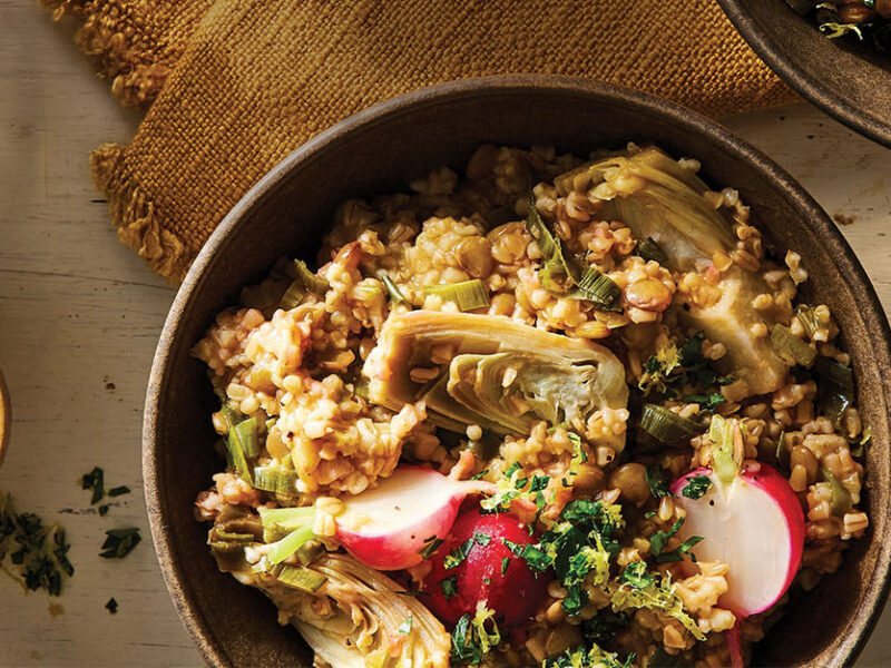 Steel-Cut Oat Risotto with Leeks and Artichokes in a brown ceramic bowl next to a small bowl of finely chopped parsley