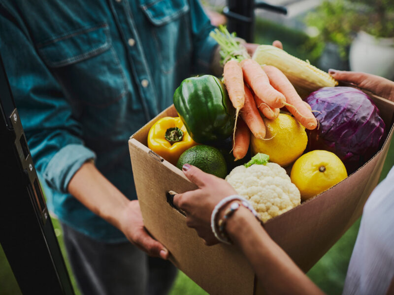 A woman hands another woman a CSA (community-supported agriculture) box full of fresh produce