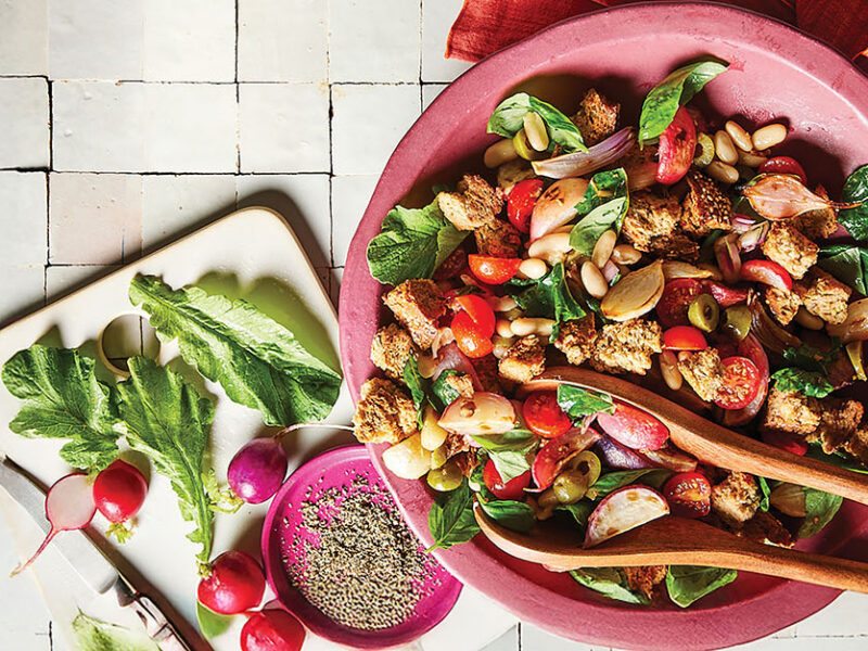 Easter Egg Radish Panzanella in a pink bowl on a white tiled background next to a plate of radishes and a small bowl of freshly ground pepper