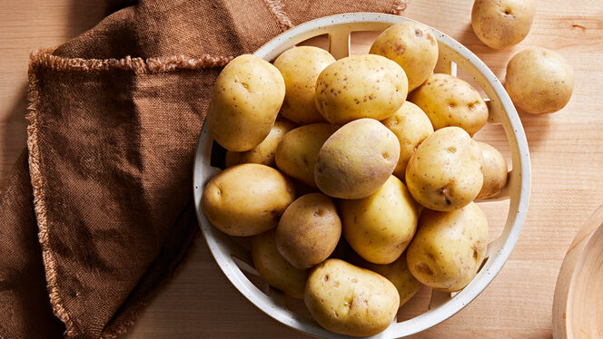 Around 20 new potatoes in a basket on a hardwood surface next to a rustic brown kitchen towel, with a couple new potatoes outsid the basket