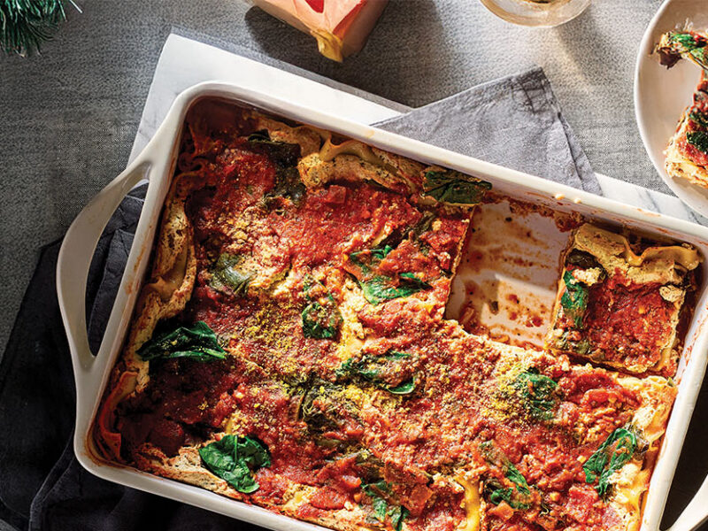 A white casserole dish of Spinach Lasagna with one slice removed and served on a white plate to the side. Set in a dimly lit grey setting.