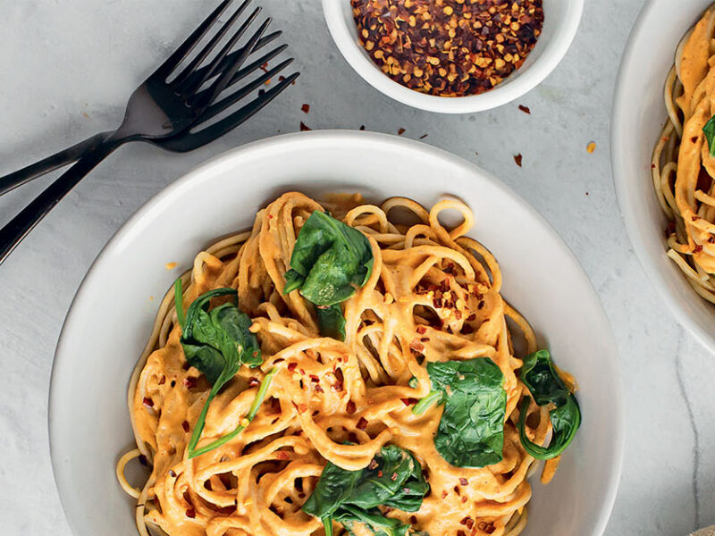 Two bowls of Brandi Doming's Harissa Almond Pasta with Spinach on a marble table with two forks and a small bowl of red pepper flakes