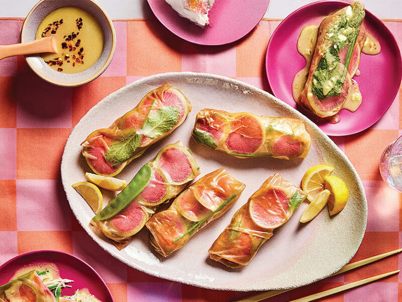 Colorful Watermelon Radish Spring Rolls on a white platter on a pink and orange checked placemat. There is a small bowl of Lemon-Ginger Dipping Sauce and two bright pink serving plates with one roll on each, smothered with sauce.
