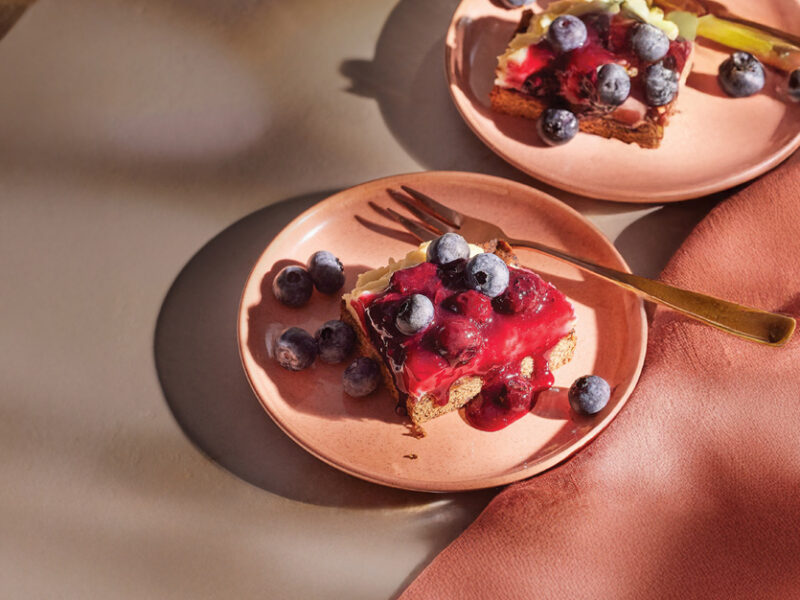 Two pale pink plates, each with a Blueberry Cheesecake Bars and garnish of fresh blueberries