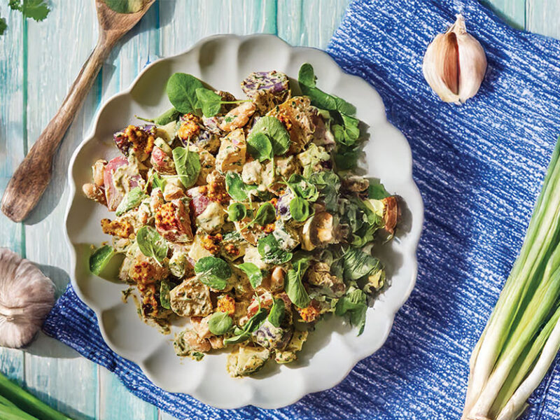 New Potato Salad with Herbed Vinaigrette on a plate on a blue-washed wooden table surrounded with a bulb of garlic, a bunch of spring onions, and a bunch of flat leaf parsley