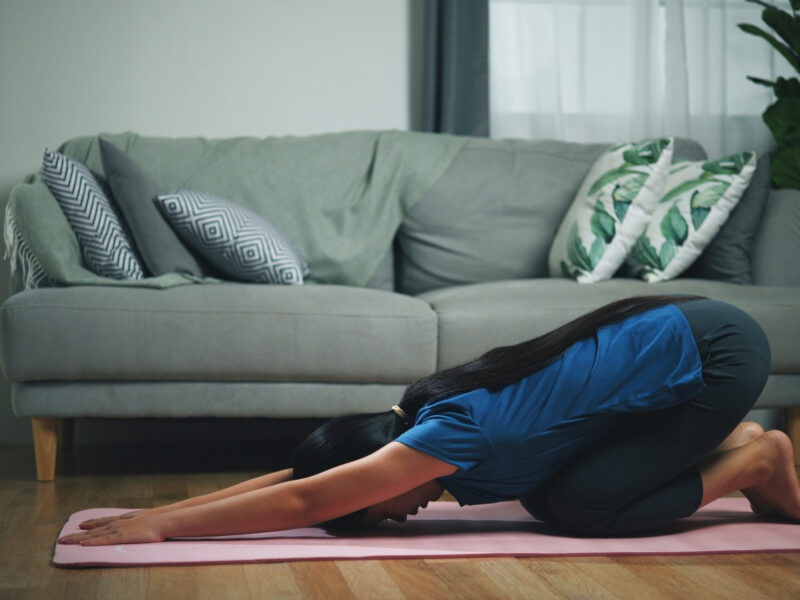 A woman in her dim living room at dusk engaged in child's pose, a restorative yoga pose to help with sleep and relaxation