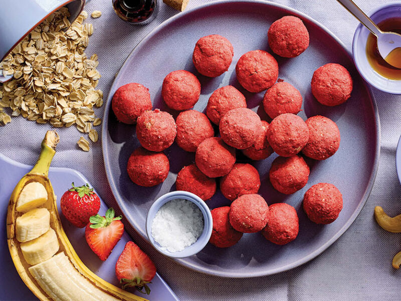 Strawberry Bliss Bites on a blue plate with small containers of cashews, rolled oats, and a banana, nearby
