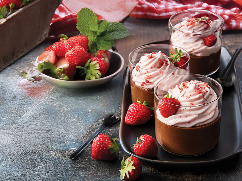 Three glass parfaits of Black Bean Chocolate Mousse with Whipped Strawberry Cream on a small tray, with fresh strawberries scattered