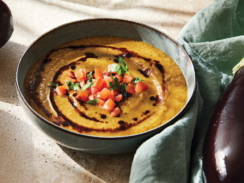 A black bowl of orange-hued soup topped with a drizzle of balsamic glaze next to a whole globe artichoke