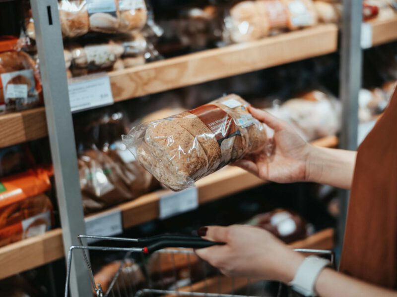A woman's hand holding up a whole grain loaf of bread at the grocery store, reading the label