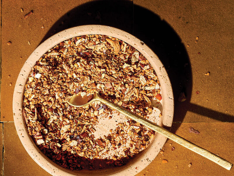 An earthy tile background and a ceramic bowl of Oil-Free Chili Crisp with a long-handled teaspoon in the bowl