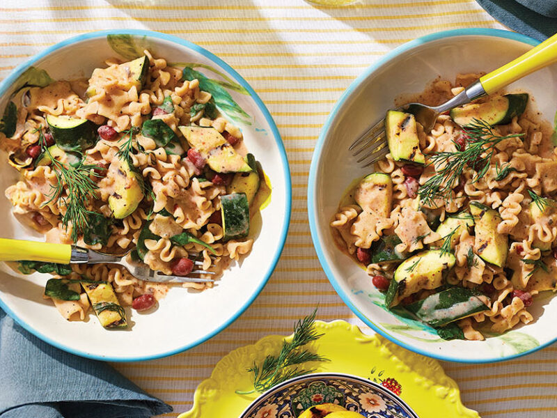 Two bowls of Charred Lemon and Zucchini Pasta with a yellow-handled fork in each and two blue napkins on a yellow and white striped table cloth