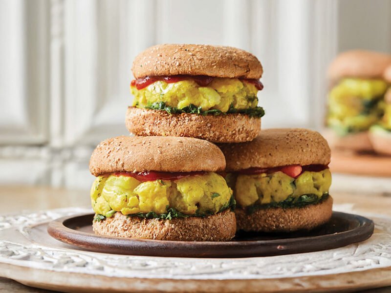Three Bombay Potato Burgers on a plate, with another plate of burgers in the background