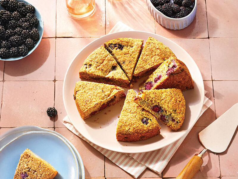 Blackberry Polenta Cake on a pink plate with one slice removed and on a nearby plate. There are two small bowls of fresh blackberries in the background