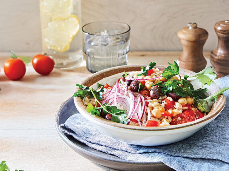 A bowl of Lemon Orzo Salad garnished with red onion and fresh parsley, with cherry tomatoes dotted on the table around the bowl