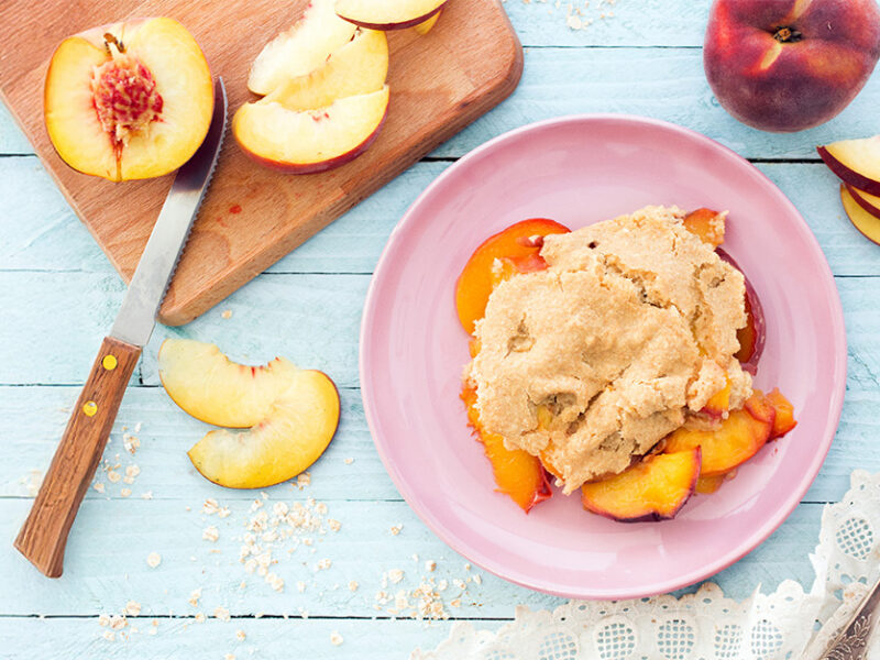 A serving of Vegan Peach Cobbler on a pink plate next to a chopping board, knife and fresh peaches, cut in half and in slices