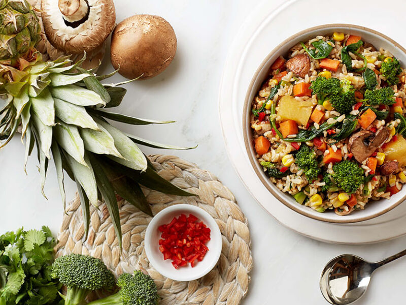 A bowl of No-Fry Fried Rice next to some raw mushrooms, broccoli, and a whole pineapple