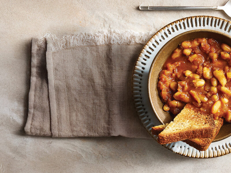 A bowl of baked beans made in a slow cooker, in a serving bowl with a slice of whole grain bread on the side