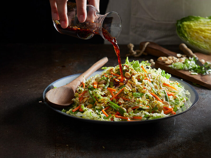 Napa Cabbage Salad in a bowl on a black counter, with a person pouring dressing over the top