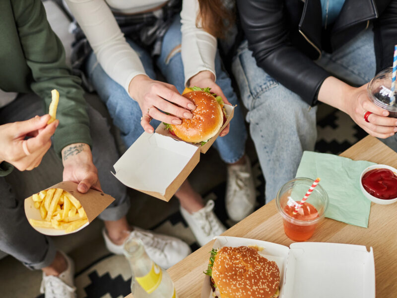 Young people sit around a coffee table eating ultraprocessed fast-food including cheeseburgers, French fries, and frozen soft drinks