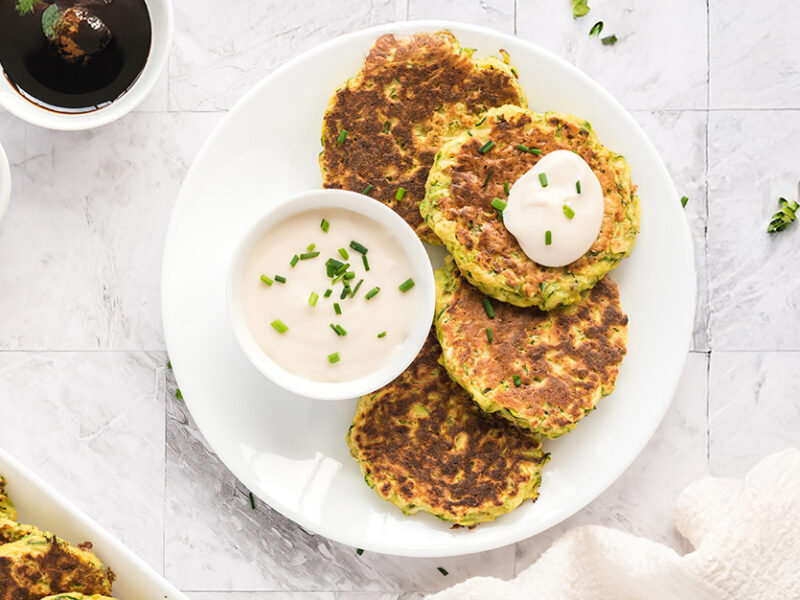 Zucchini Fritters on a white plate with a small white bowl of sauce, next to a larger serving plate of fritters