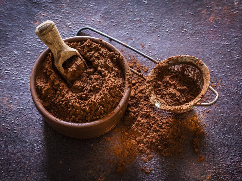Brown bowl filled with cocoa powder shot on rustic table. Beside the bowl is a sieve also with cocoa powder.