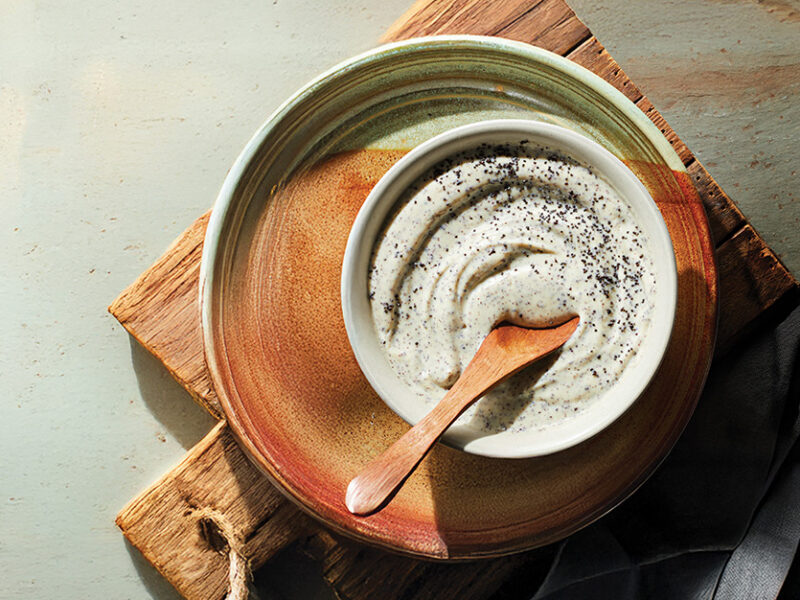 A rustic bowl of creamy poppy seed dressing on a n earthy handmade ceramic plate on a wooden chopping board
