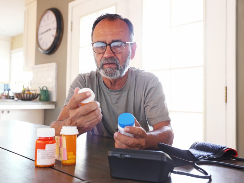 A senior citizen man sits at his dining room table sorting through bottles of prescription medications as sunlight filters in through the window behind him bathing the room with a soft glow of light. A blood pressure machine rests on the table in front of him.