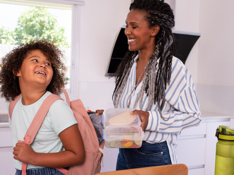 A mother stands behind her daughter in the kitchen ready to put a bag of snacks into her daughters backpack. Both are smiling and happy.