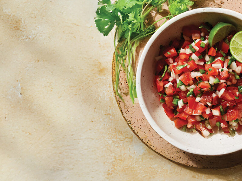 Pico de Gallo in a pale pink bowl on a brown ceramic plate, with cilantro on the side, and lime wedges in the bowl