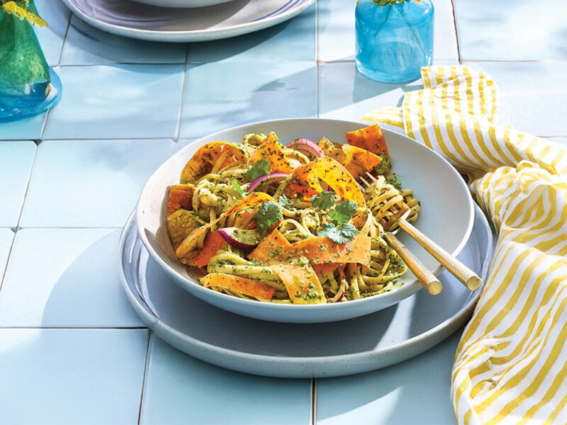 A bowl of Fettuccine with Spinach Pesto and Carrot Ribbons in a pale blue bowl on a blue plate on pale blue tiles with a yellow and white striped unfolded napkin next to the plate